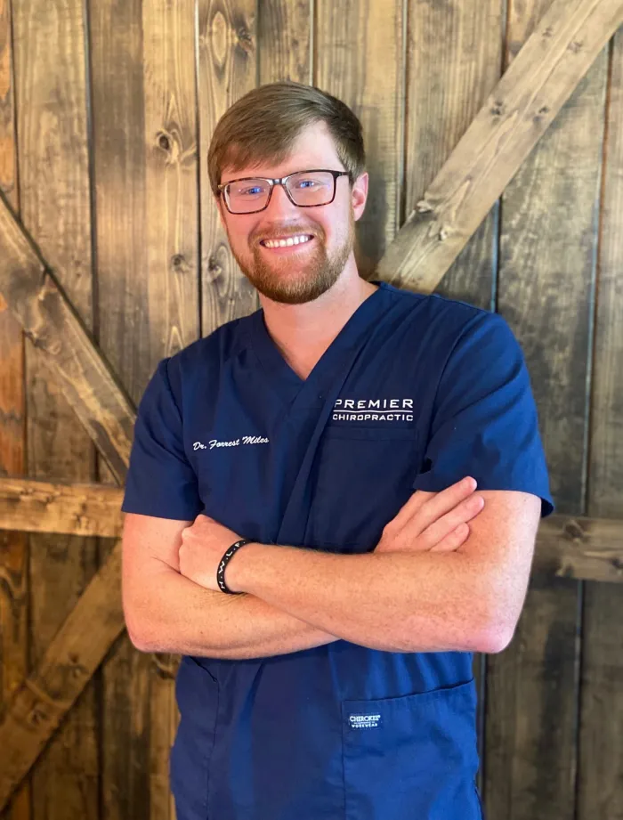 Dr. Forrest Miles in glasses and navy scrubs, arms crossed, in front of a barn wood wall