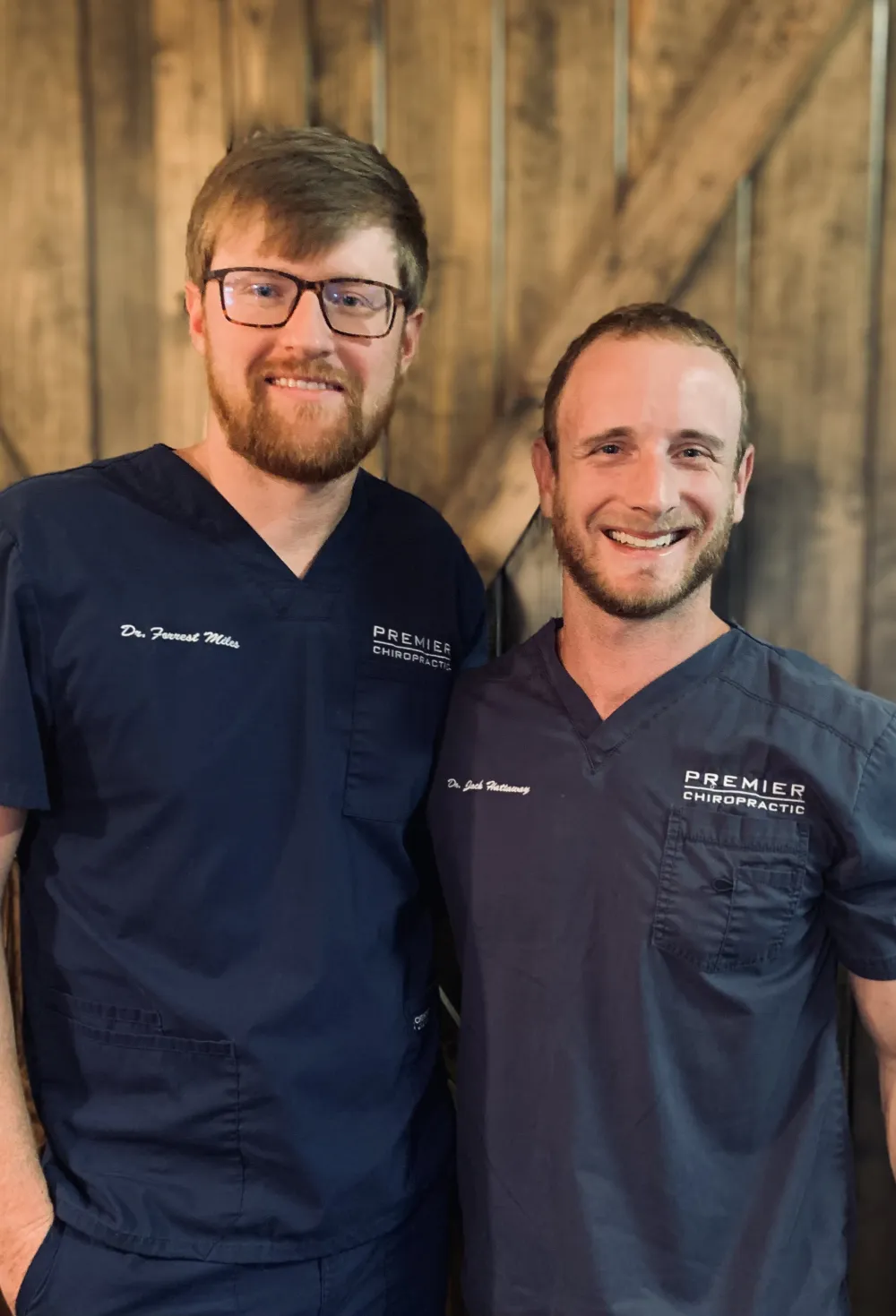 Dr. Forrest Miles and Dr. Jack Hattaway smiling side by side in navy Premier Chiropractic scrubs in front of a barn wood wall