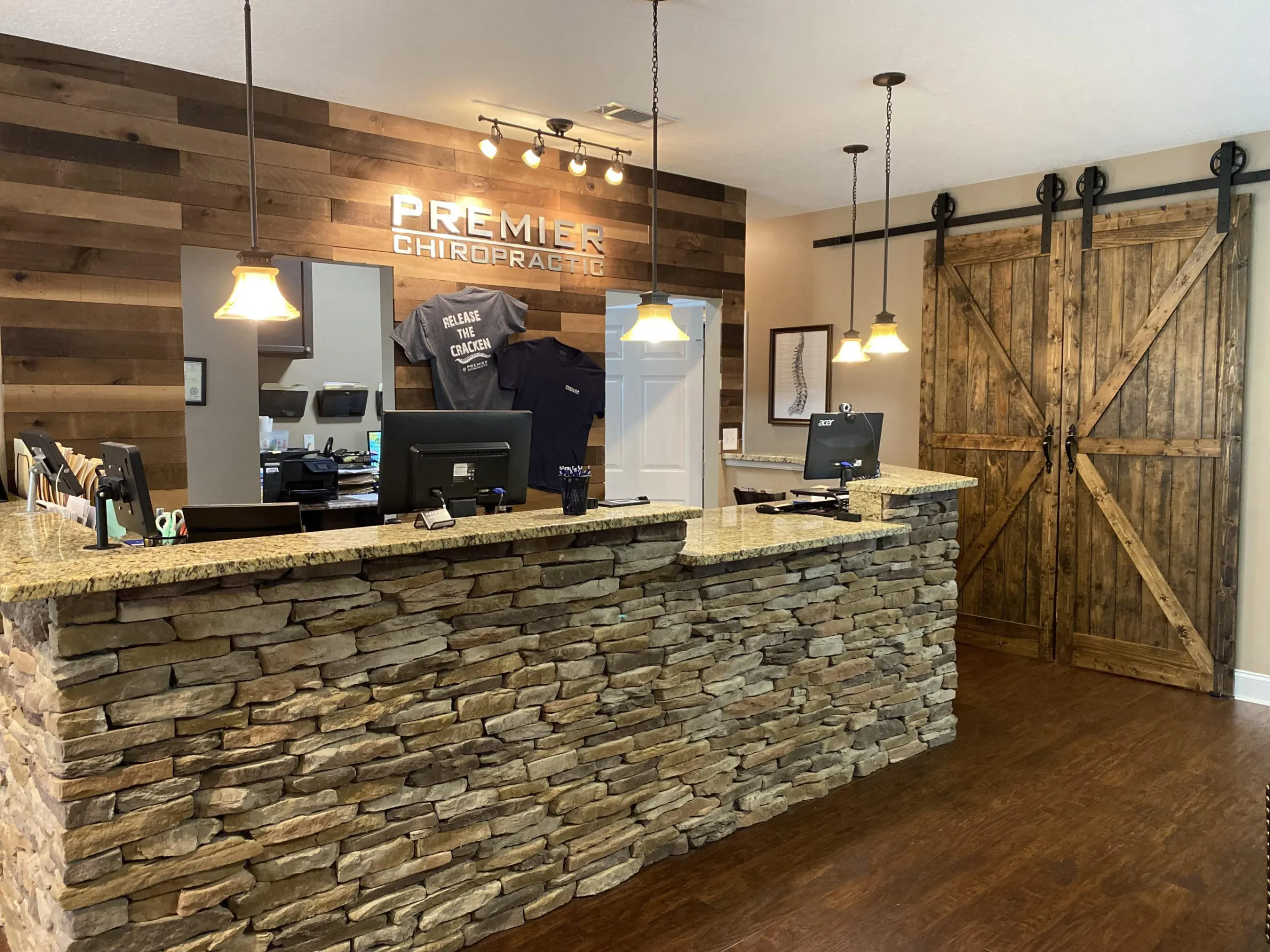 The Premier Chiropractic front desk: a stacked stone counter, a wood plank wall with the Premier Chiropractic sign, pendant lights and sliding barn doors