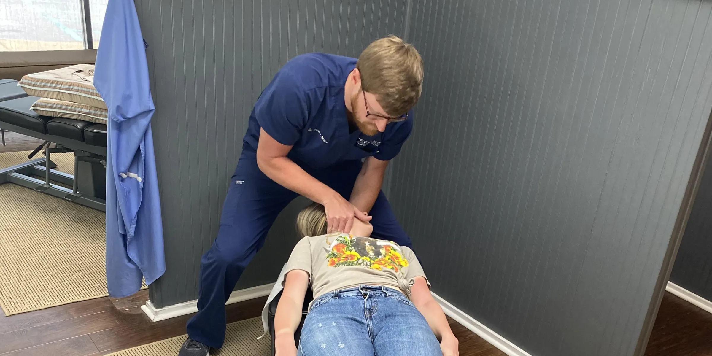 A doctor with his hands at the neck and shoulders of a patient lying face up on an adjustment table, beside a grey partition